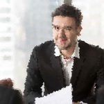 Man in a dark suit speaks to colleagues during a business meeting, holding papers